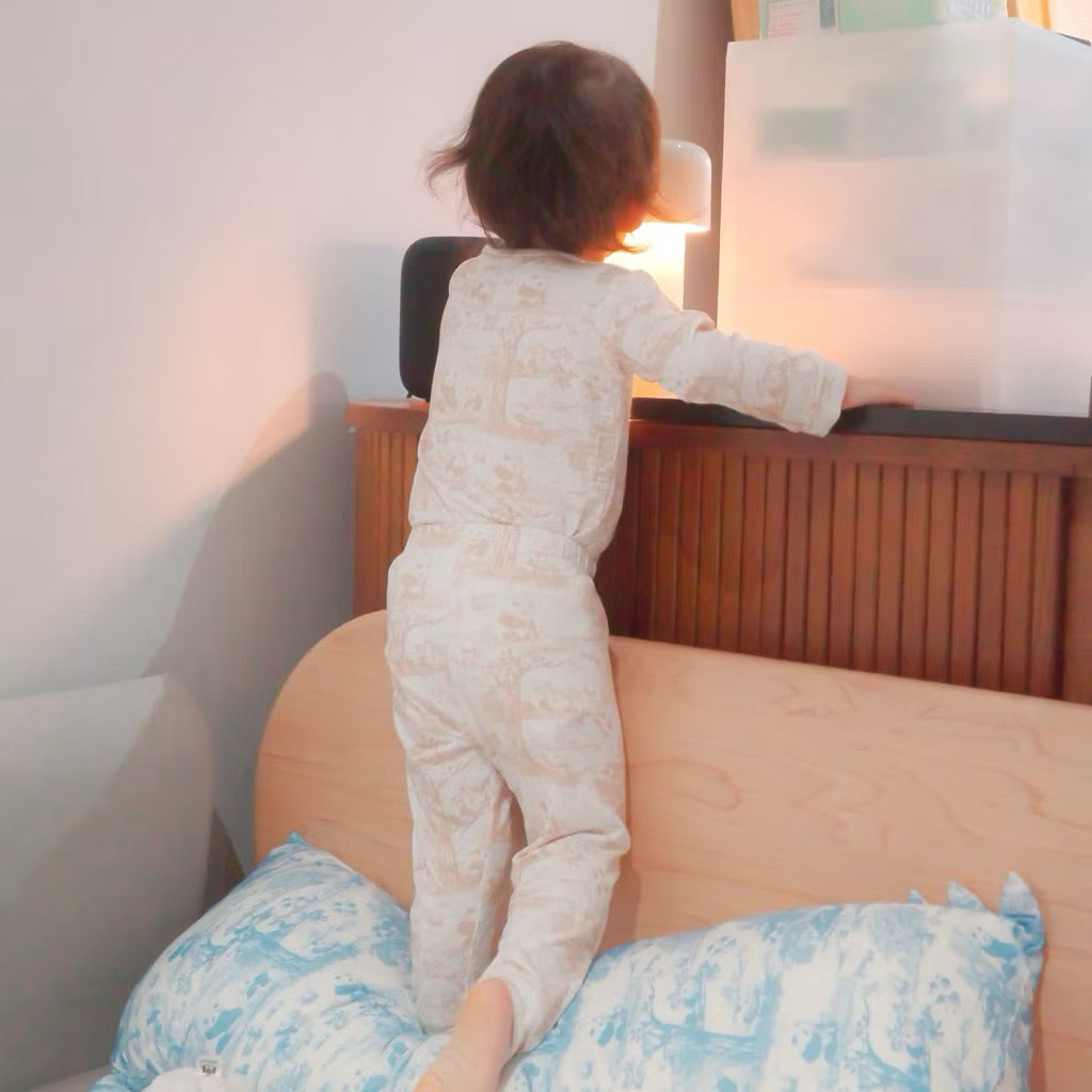 Child wearing bamboo kids pyjamas standing on a bed beside a nightstand with a lamp — soft and eczema-friendly sleepwear in Singapore.