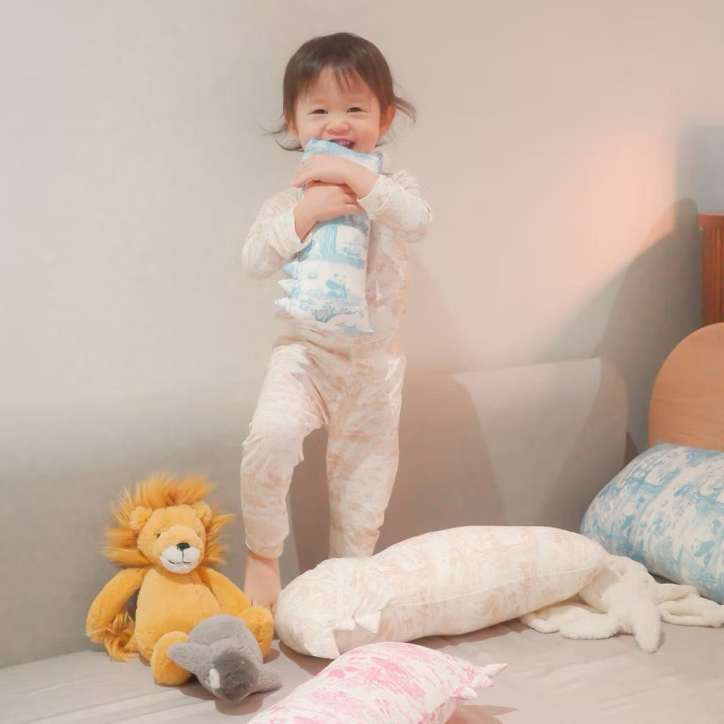 Child in bamboo kids pyjamas holding a small blue bamboo pillow on a bed surrounded by stuffed animals and pillows — cooling, eczema-friendly sleepwear and bedding