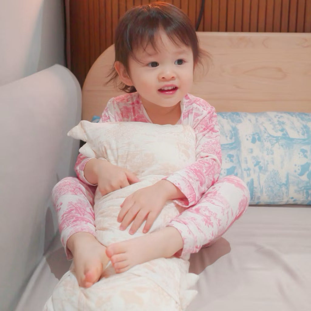 Child in pink bamboo pyjamas sitting on a bed while holding a white pillow with a beige pattern — soft, cooling, eczema-friendly sleepwear and bedding.
