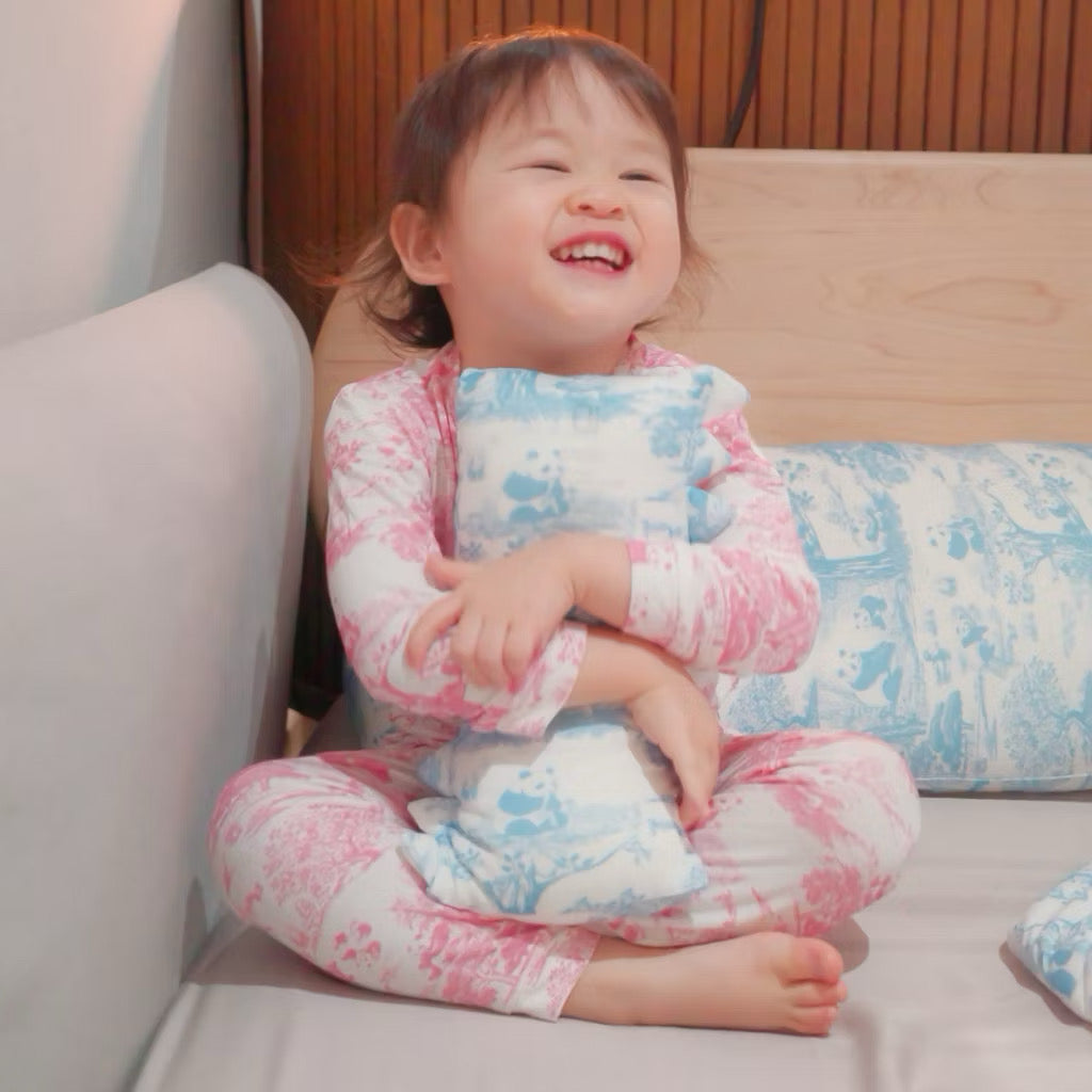 Child in pink bamboo pyjamas hugging a blue bamboo pillow while sitting on a bed with multiple pillows — cooling, eczema-friendly sleepwear and bedding.