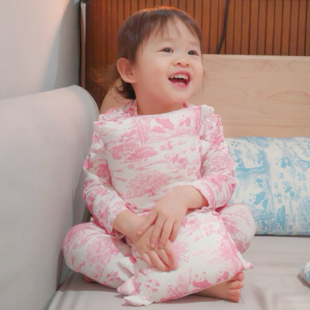 Child in pink bamboo pyjamas sitting on a bed and hugging a pink bamboo pillow against a neutral background — soft, cooling, eczema-friendly sleepwear and bedding.