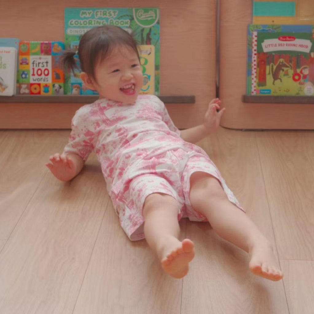 Child wearing a pink short sleeve 2-piece bamboo kids pyjamas set lying on a wooden floor with books in the background — soft and eczema-friendly sleepwear.