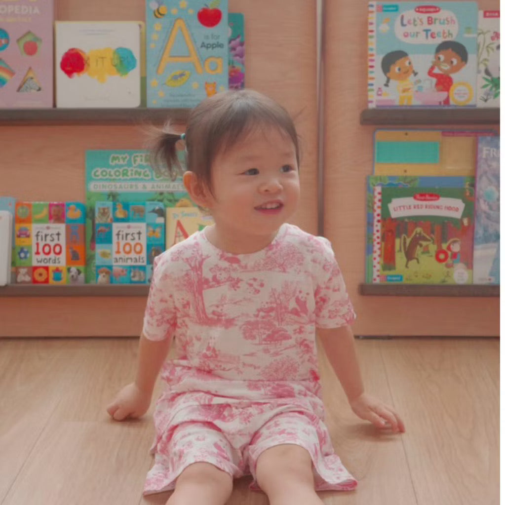 Child wearing a pink short sleeve 2-piece bamboo kids pyjamas set sitting on a wooden floor with children’s books in the background — cooling, eczema-friendly sleepwear.