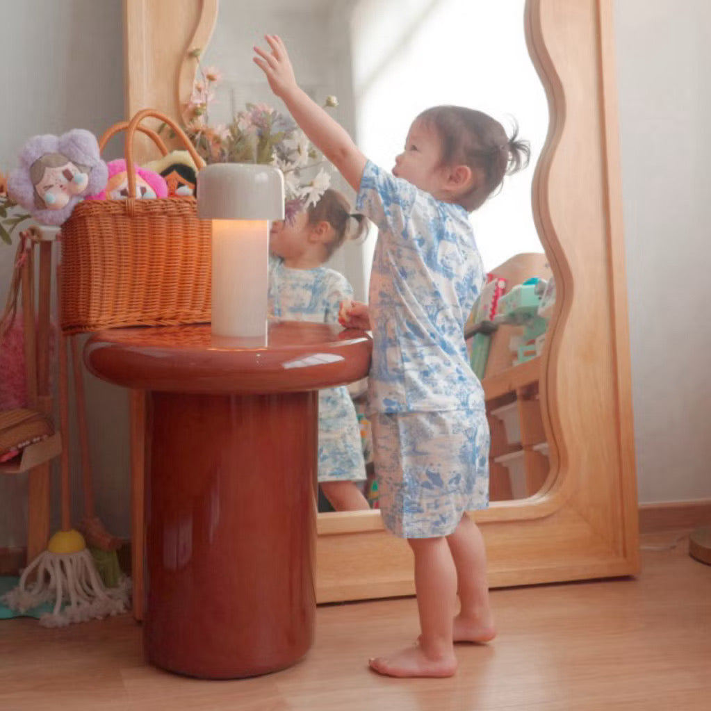 Child wearing a pink short sleeve 2-piece bamboo kids pyjamas set standing beside a small table with a lamp and looking into a mirror — cooling and eczema-friendly sleepwear in Singapore.